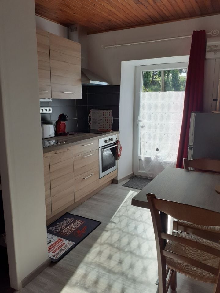 The kitchen area features light wood cabinetry and modern appliances, including an electric oven and refrigerator. A small dining table with wooden chairs is positioned near a window covered by a sheer curtain, allowing natural light to brighten the space.