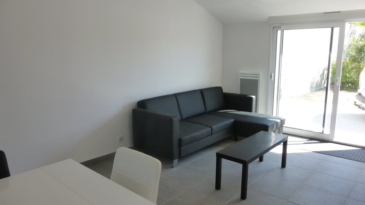 A modern living area features a sleek black sofa with an extended chaise lounge, positioned opposite a simple coffee table. Large glass doors allow natural light to fill the room, with a view of greenery visible outside. The space is adorned with neutral-toned walls and tiled flooring.