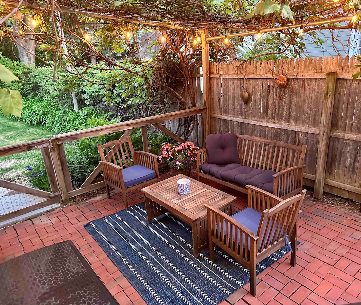 An outdoor patio is adorned with string lights and surrounded by a wooden fence. Wooden furniture includes a sofa and two chairs, arranged around a coffee table on a textured rug. Potted flowers add a touch of color to the space, complemented by lush greenery in the background.