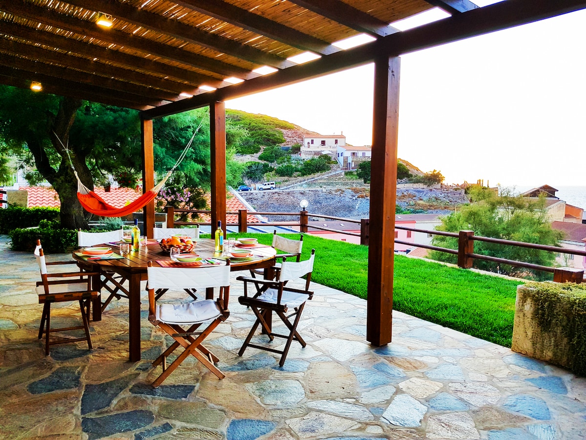 An outdoor patio is featured with a large dining table surrounded by wooden chairs. A hammock is seen to the side, and the setting includes a grassy area, stone flooring, and a view of the hillside and nearby buildings. Soft lighting enhances the tranquil ambiance.