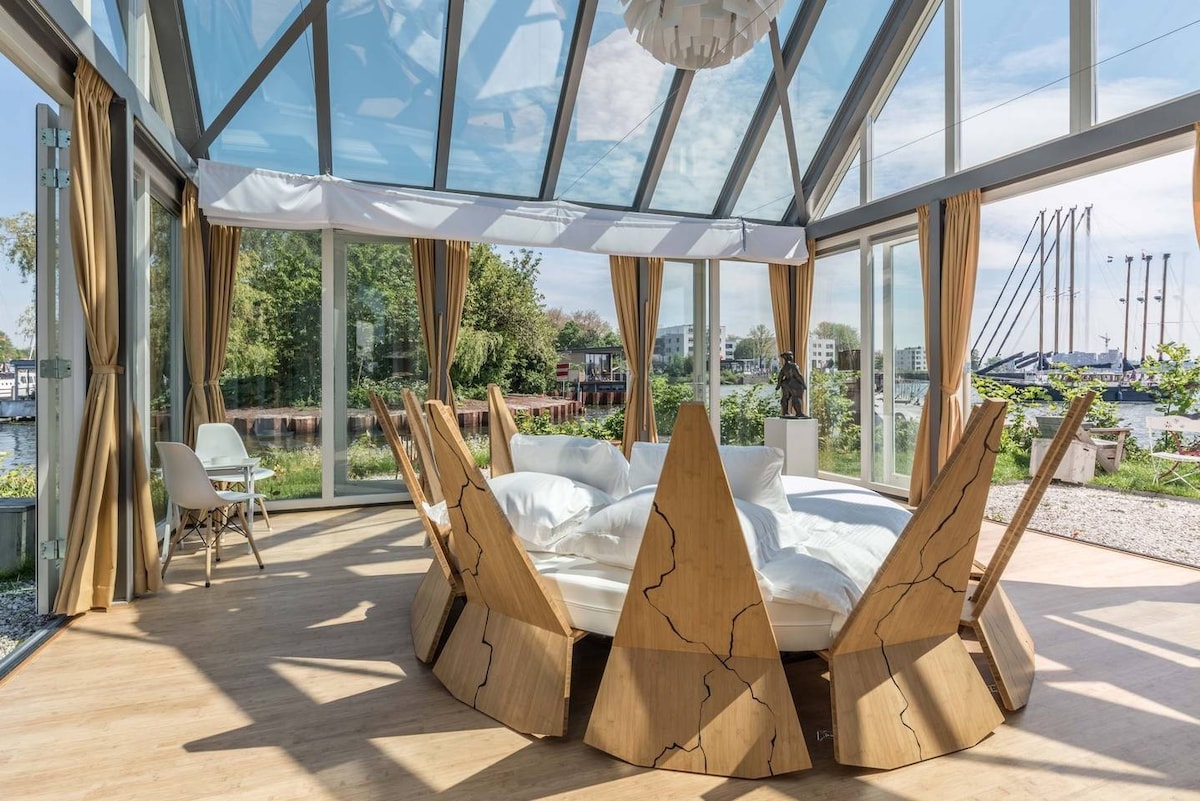 A spacious conservatory features a uniquely designed round bed surrounded by wooden elements, resembling petals. Large glass panels invite natural light and provide views of the landscape outside. Light curtains frame the windows, and contemporary seating is arranged for relaxation.