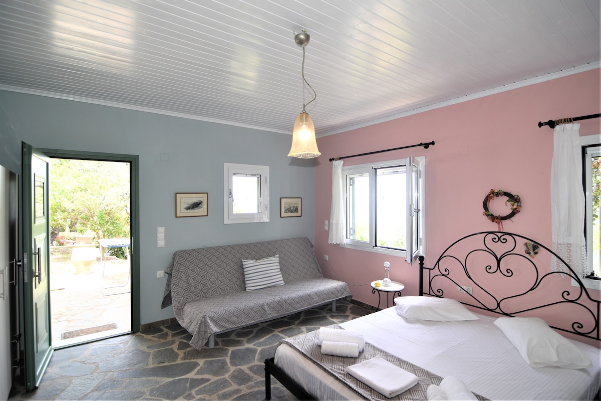 The studio features a blend of soft pink and blue walls, creating a light and airy atmosphere. A spacious bed with white linens is complemented by a comfortable sofa. Natural light floods in through several windows, while a door opens to a scenic outdoor view.