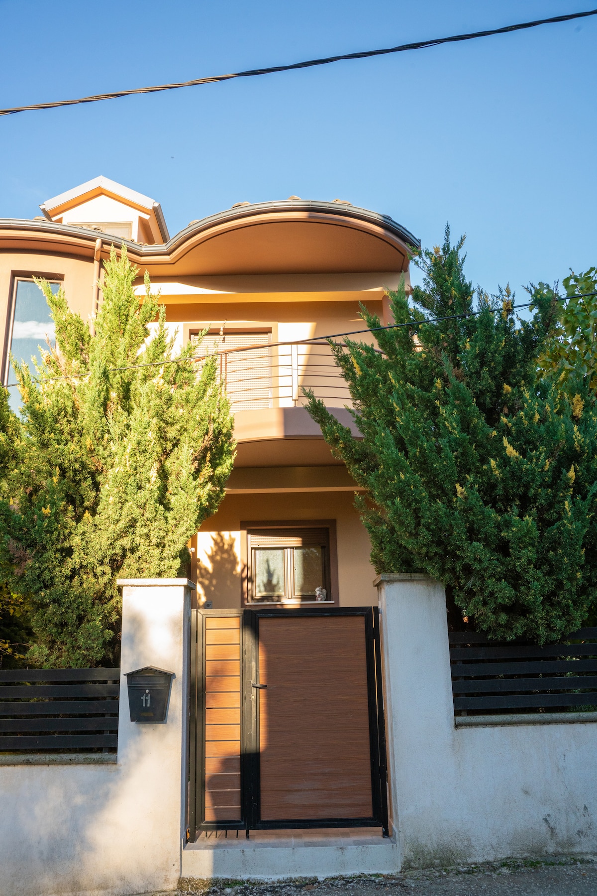 The entrance of the house features a modern security door framed by tall greenery. The structure is two stories high, with a balcony visible on the upper level. A clear blue sky complements the warm tones of the exterior walls.