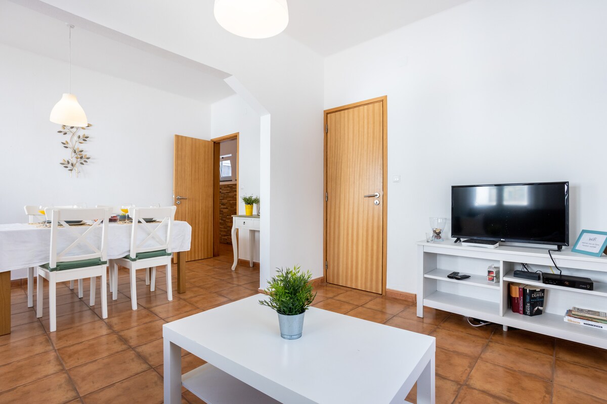 A bright living area features a white coffee table and a small potted plant. A TV is positioned on a low shelf, while a dining table with six chairs is visible in the background. Two doors provide access to other rooms in the space.