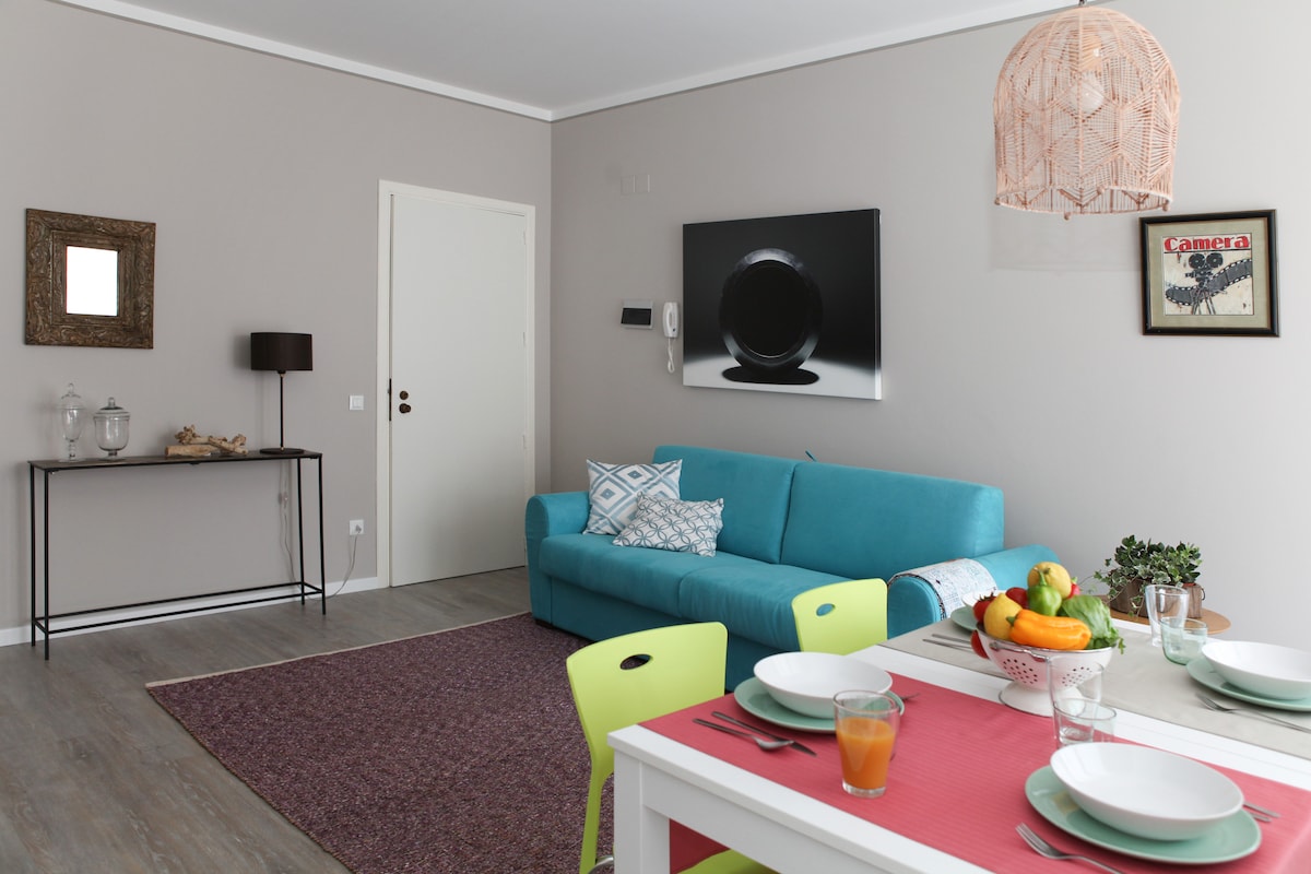 A bright living area features a turquoise sofa, light wood flooring, and a dining table set for four. A colorful fruit bowl is visible on the table, while a wall art piece adds a modern touch. Natural light enters through a nearby window.