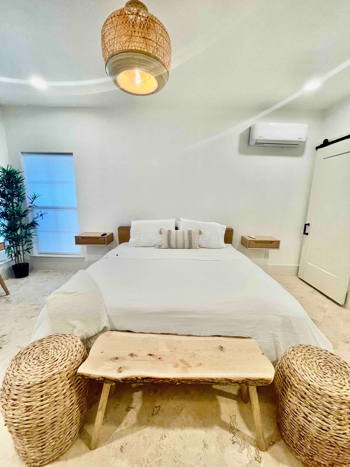 The bedroom features a spacious bed with neutral bedding and decorative pillows, complemented by two mounted nightstands. A wooden bench stands at the foot of the bed, while a woven pendant light hangs overhead. Natural light enters through a nearby window, illuminated by a soft glow.