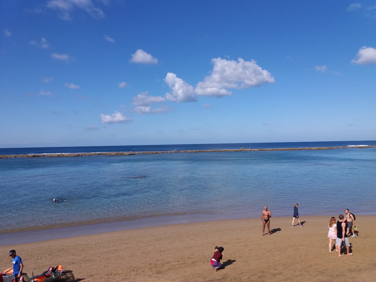 A sandy beach is depicted, featuring gentle waves lapping at the shore. Various individuals can be seen enjoying the sun and the water, with a clear blue sky overhead and scattered clouds. The tranquil setting invites relaxation and leisure activities.