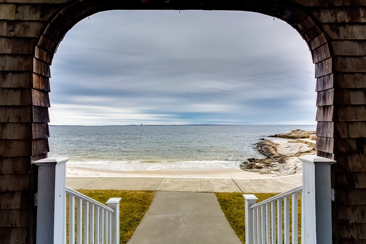 A view of the beach and ocean is framed by an arched entrance. White railings and a concrete path lead down to the sandy shore, where gentle waves meet the land. The sky displays a gradient of soft grey and blue, enhancing the serene atmosphere.