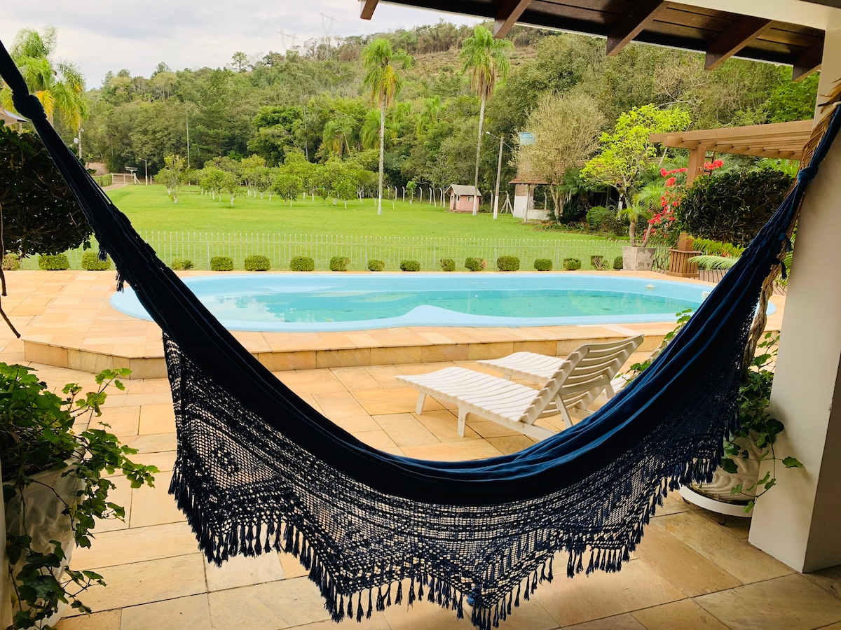A hammock is positioned in the foreground, framing a view of a swimming pool and a landscaped yard. The pool is surrounded by a tiled patio, with a sun lounger nearby. Lush greenery from trees and plants creates a serene backdrop.