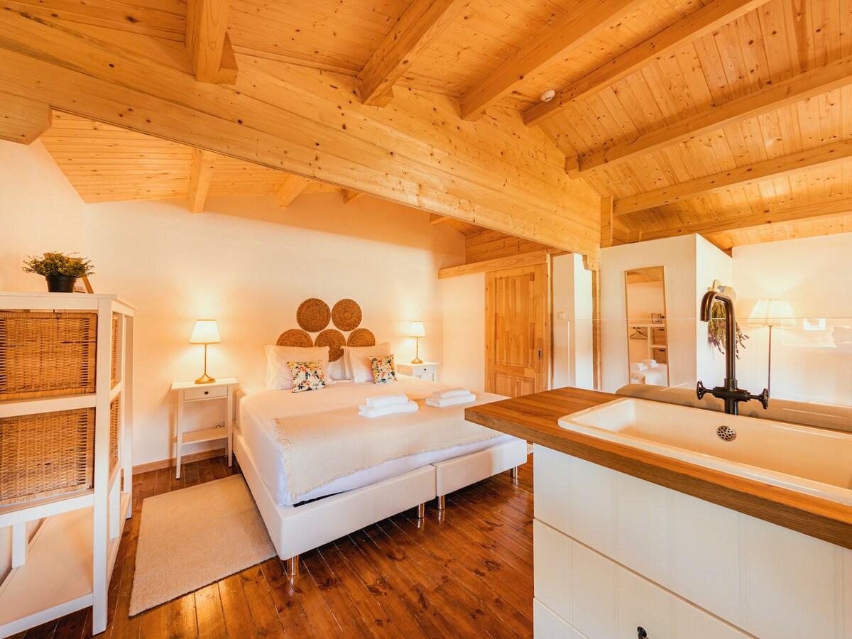 A bright sleeping area is characterized by wooden beams overhead and warm lighting from two bedside lamps. A spacious bed, adorned with white linens and decorative pillows, is positioned against a textured wall. A small sink is visible in the foreground, enhancing the room's functionality.