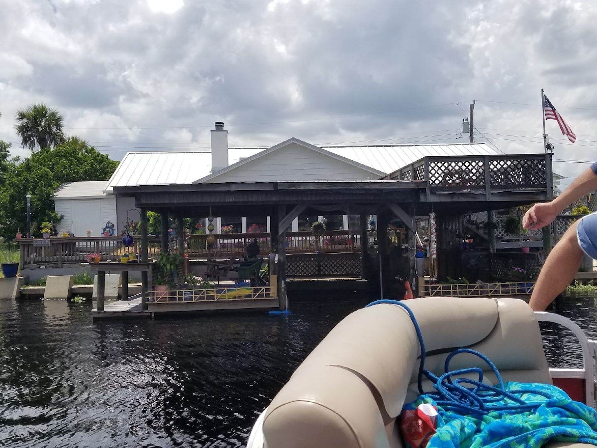 The image captures a view of the lakefront property featuring a two-story deck with shaded areas beneath. A boat dock and ramp can be seen leading into the water. The house is partially obscured by greenery, and a flag flies from a nearby pole.