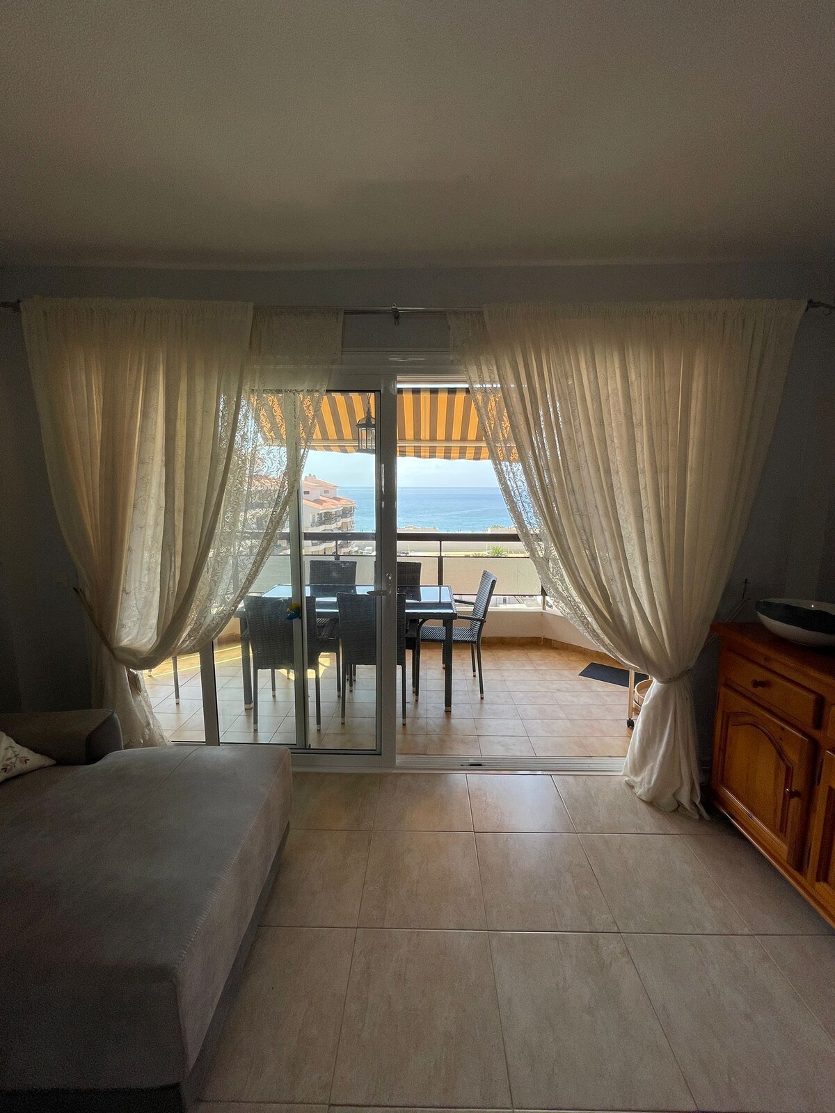 A spacious living area features large glass doors leading to a terrace, allowing natural light to fill the room. Sheer curtains frame the entrance, softly diffusing the sunlight. Outside, a dining set for six is visible on the terrace, with views of the sea in the background.