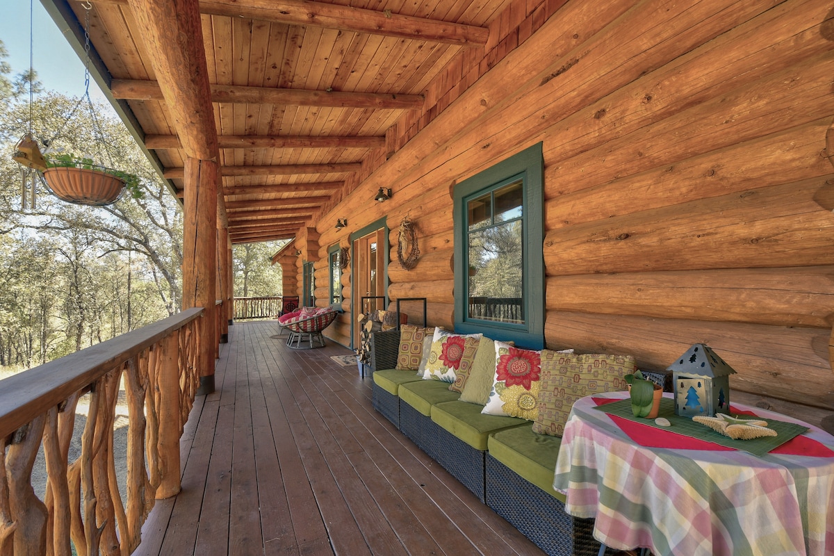 A spacious wrap-around deck is showcased, featuring a cozy seating area with bright cushions and a patterned tablecloth. The natural wood logs of the house complement the outdoor setting, while hanging plants add a touch of greenery. Sunlight filters through the trees, creating a serene atmosphere.