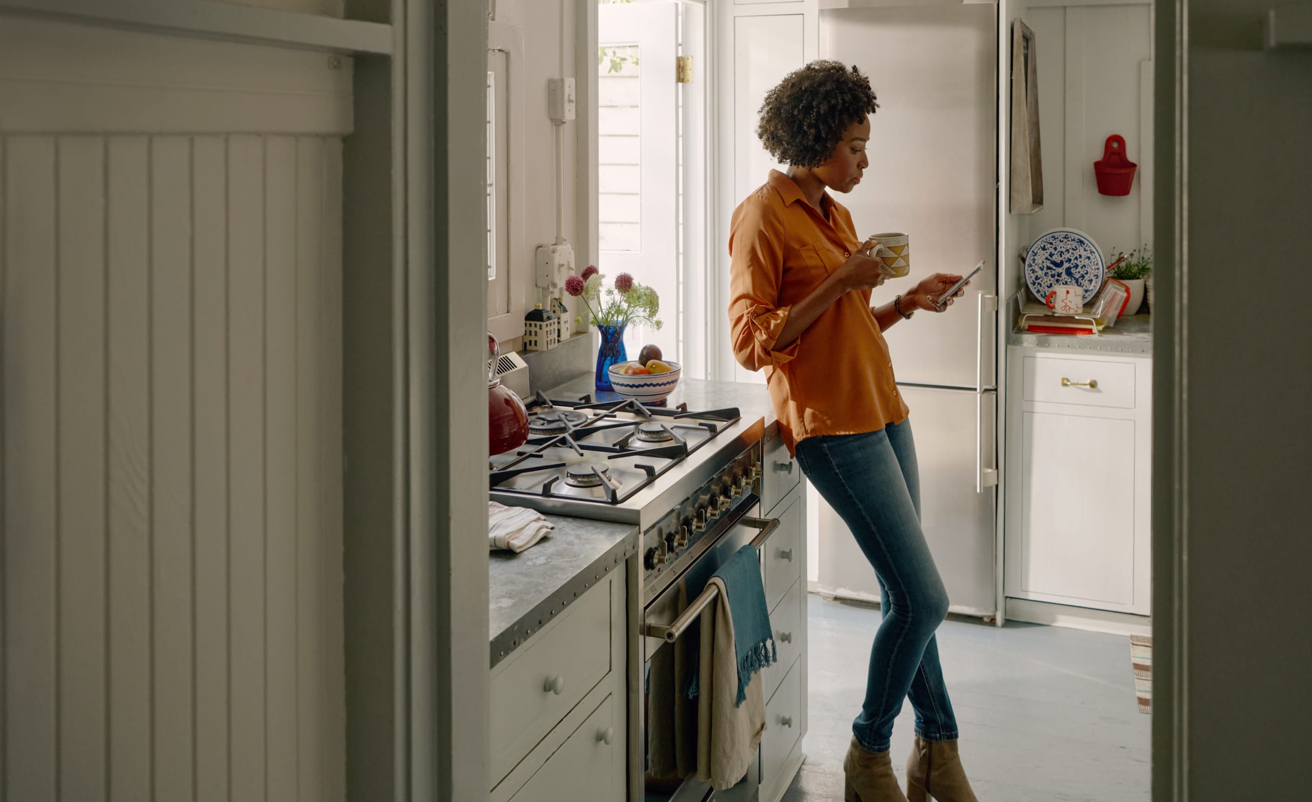 A person leans against the worktop in a kitchen, holding a mug and reading something on their mobile phone.