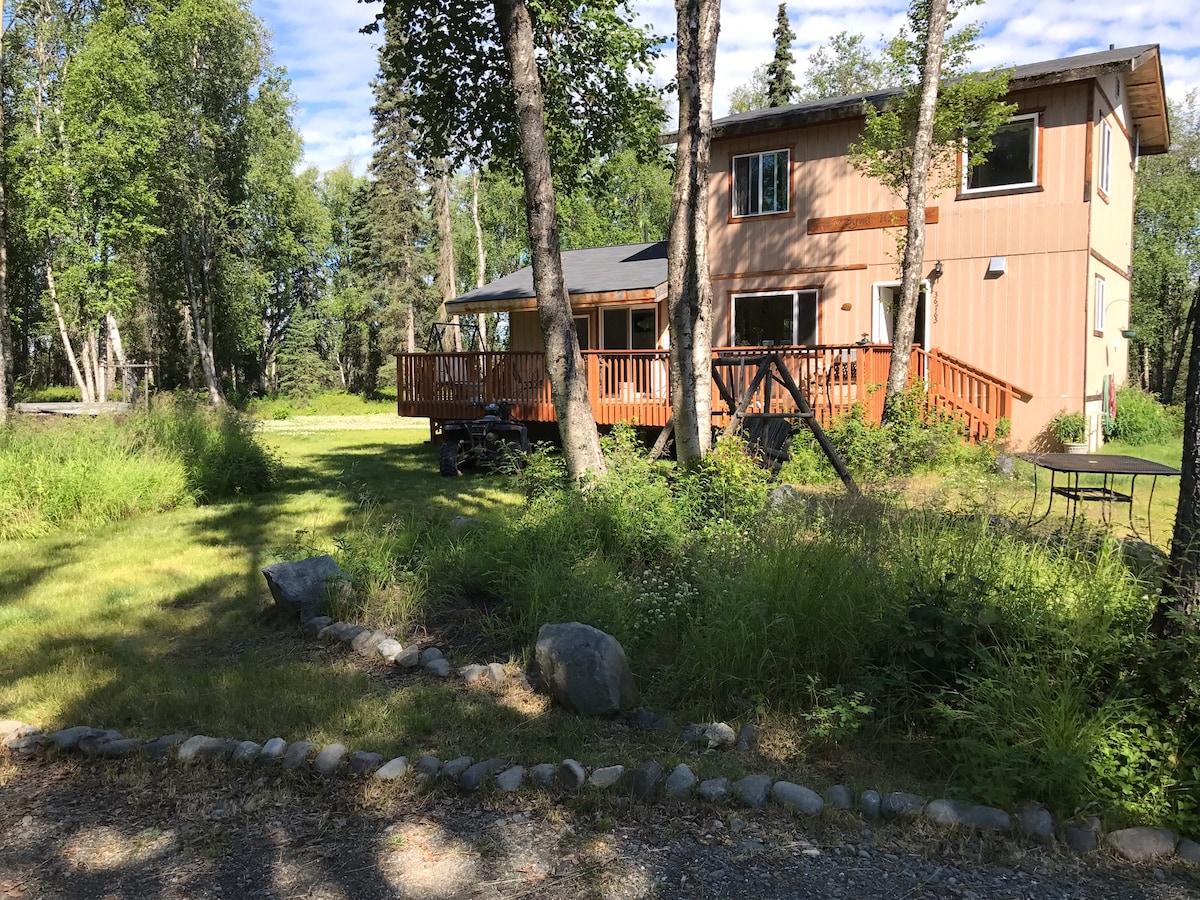 A two-story house is surrounded by trees in a natural setting. A wooden deck is visible, with seating and a swing set in the yard. Lush grass and small stones create a pathway leading to the entrance, inviting relaxation in a quiet environment.