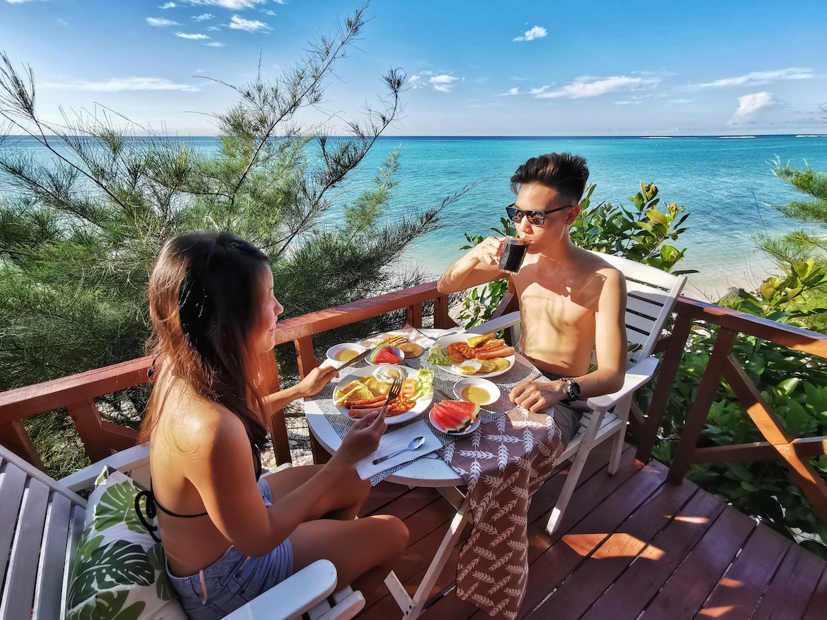 A wooden deck offers a dining setup with two chairs facing the ocean. A table is set with an assortment of meals, surrounded by lush greenery. The clear blue water stretches into the distance under a bright sky, creating a serene atmosphere for dining.