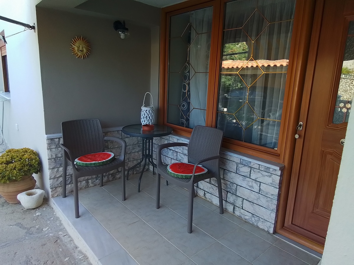 A small outdoor seating area is presented, featuring two chairs with round cushions and a glass-top table. A decorative wall element adds character, while natural light flows in through the large window, creating a cozy and inviting space.