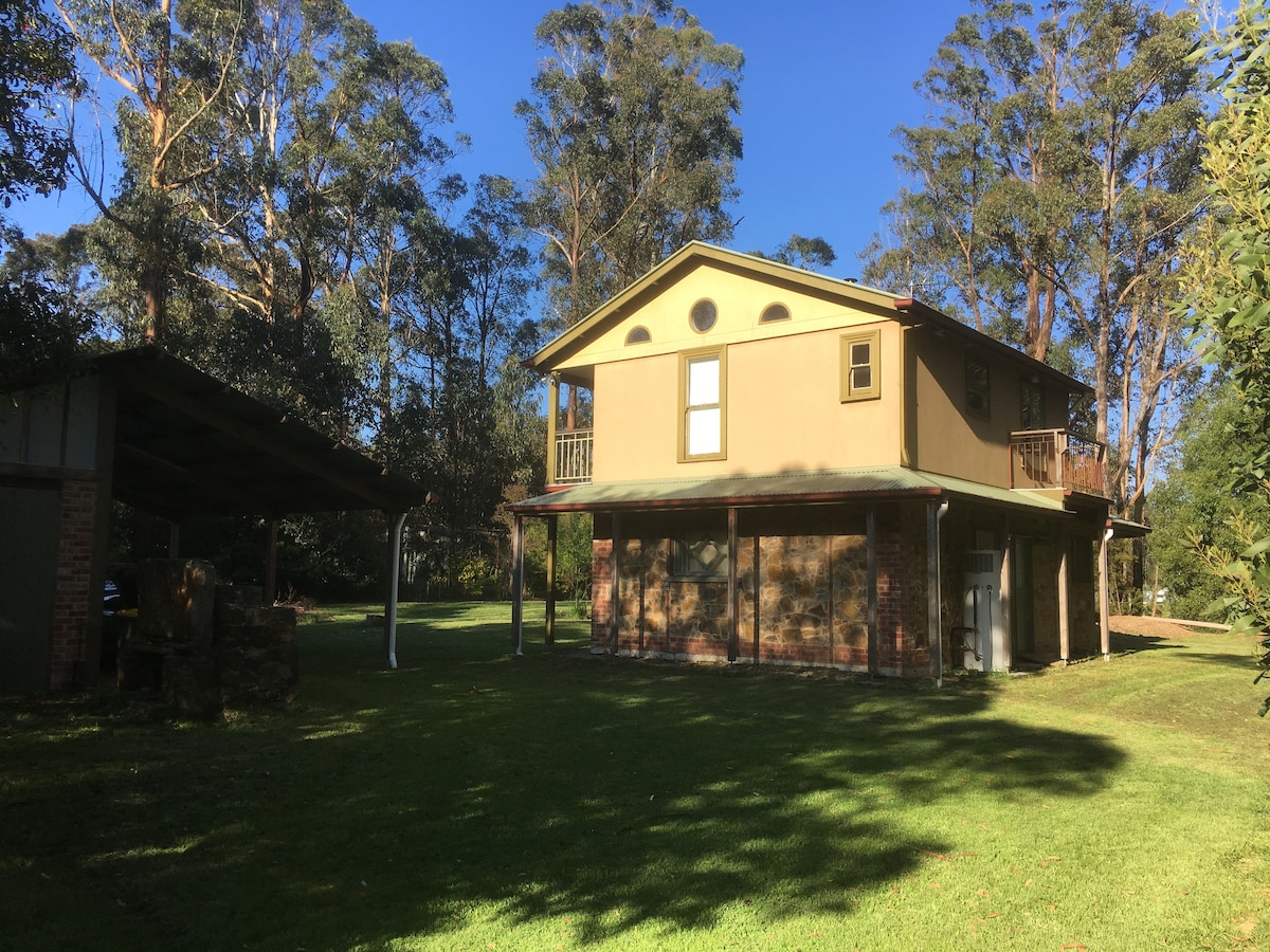 A two-story stone cottage sits surrounded by lush greenery, with tall trees providing shade. The exterior features a combination of stone and rendered walls, and spacious glass windows allow natural light. A covered outdoor area is visible to the left, enhancing the inviting landscape.