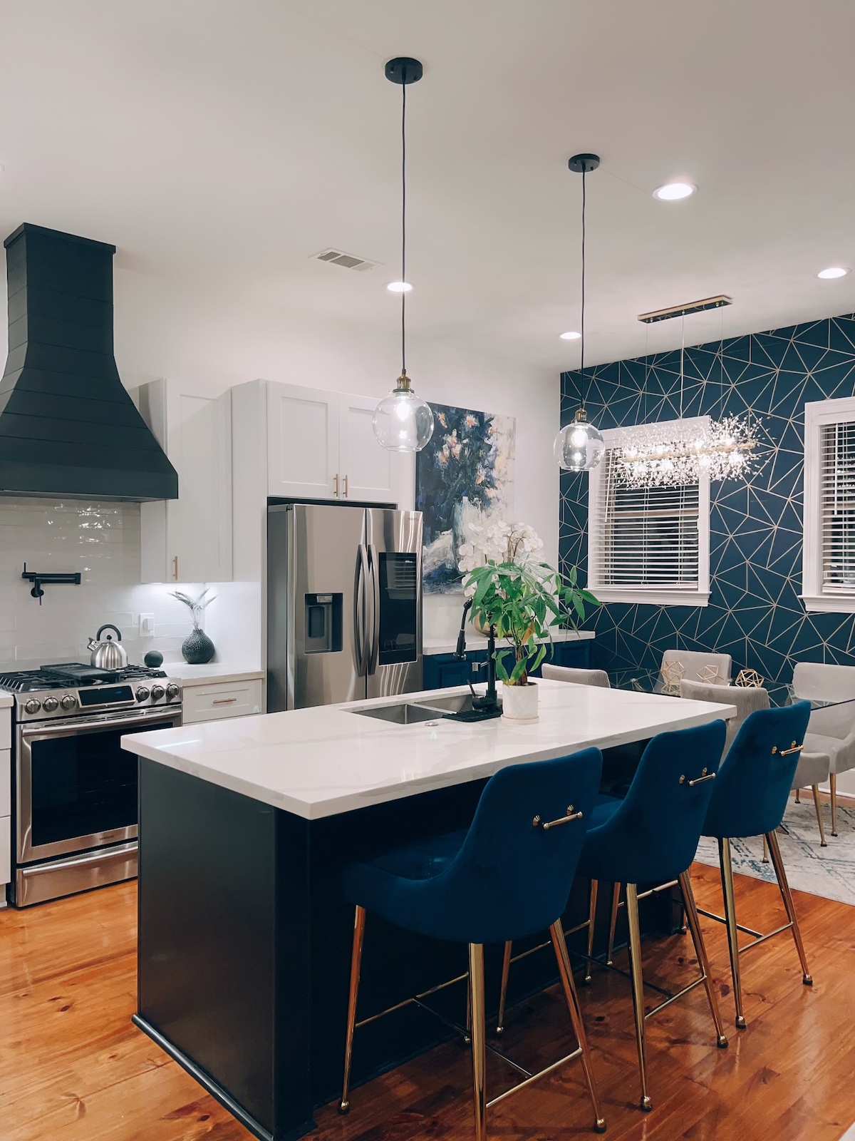 A modern kitchen features a large central island with four blue upholstered bar stools. Sleek stainless steel appliances are integrated, alongside a stylish vent hood. Soft lighting illuminates the dark blue geometric accent wall, complemented by a colorful wall art piece and large windows allowing natural light to enter.