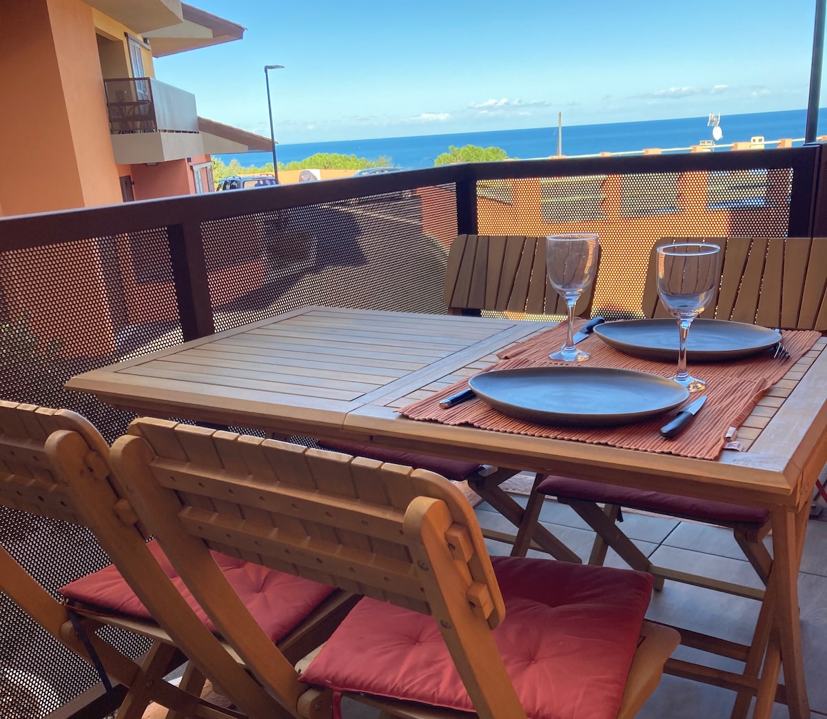 A wooden table is set on a terrace, accompanied by four folding chairs. Table settings include plates and glasses, highlighting a space ideal for dining outdoors. A scenic view of the sea is visible in the background, framed by a clear blue sky.