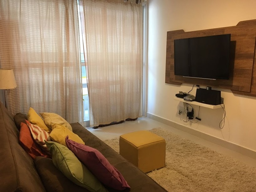 A comfortable living area features a soft, neutral carpet and a brown sofa adorned with colorful cushions. Natural light filters through sheer curtains covering a large window. A wall-mounted television is positioned above a small media console, offering a cozy entertainment space.