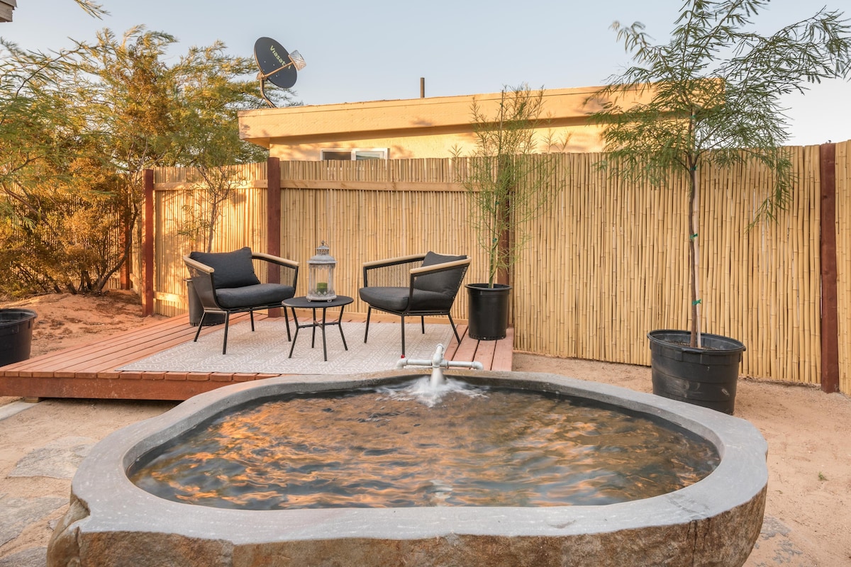 A private rock soaking tub filled with mineral-rich hot springs water is centrally featured. Two black lounge chairs are positioned beside it on a wooden deck, enhancing the outdoor space. A natural bamboo fence provides a backdrop, with potted plants adding greenery.