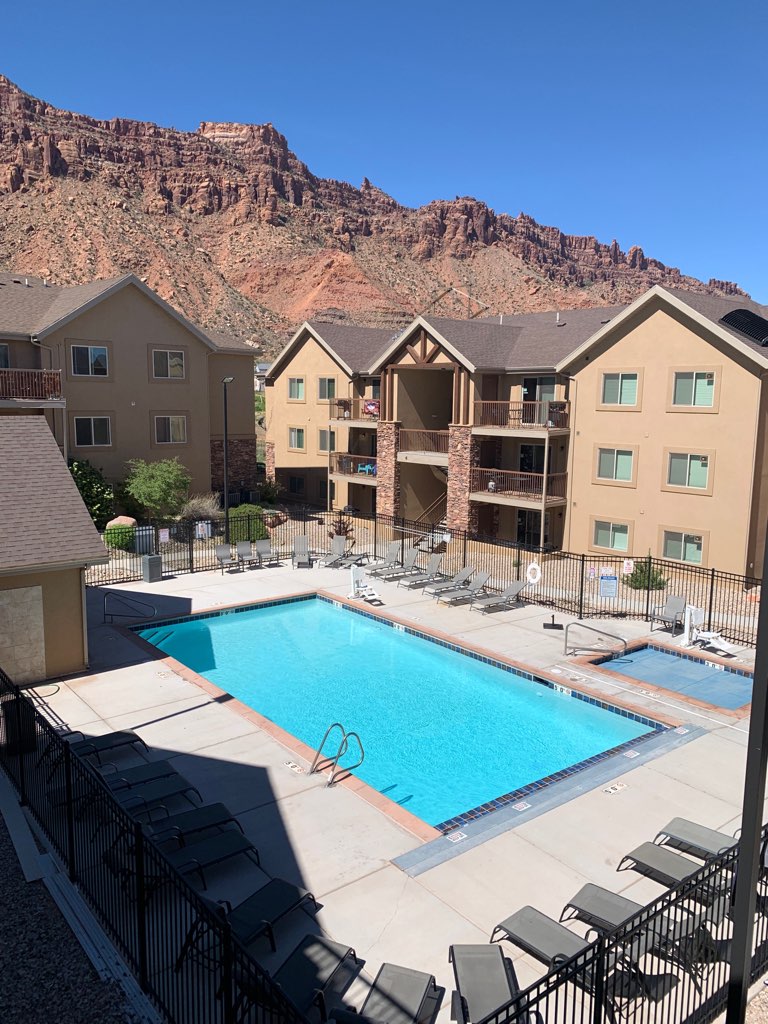 A large outdoor pool surrounded by a spacious deck features comfortable lounge chairs and is situated within a gated area. The backdrop displays impressive red rock formations under a clear blue sky, enhancing the natural beauty of the location.