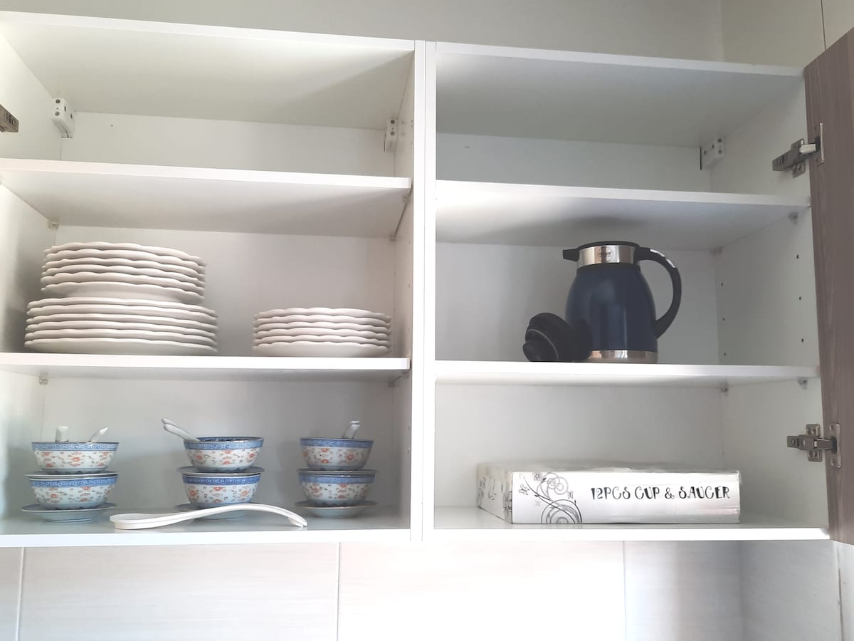 A cupboard is filled with various dining essentials. Stacked plates are neatly arranged on the top shelf, while bowls rest on the bottom shelf beside a kettle. A white box labeled '12PCS CUP & SAUCER' is positioned at the front, offering organized storage.