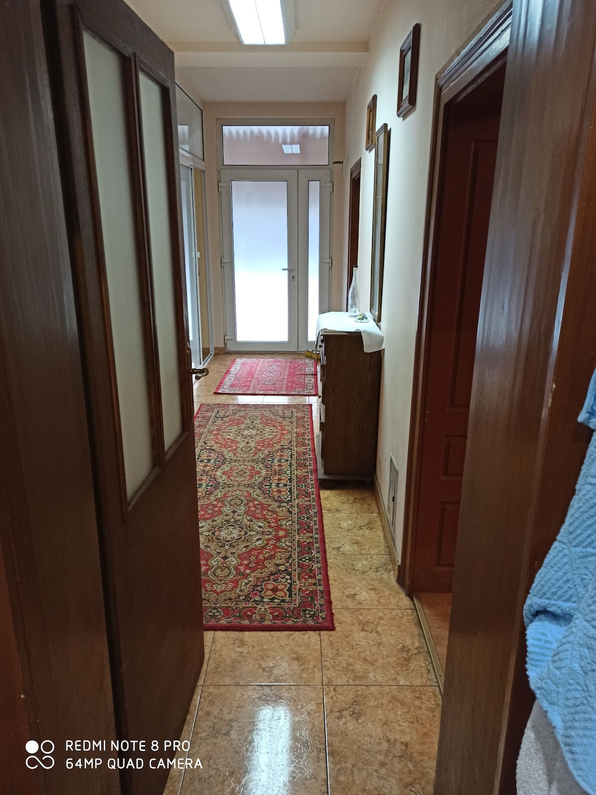 A hallway is shown with glossy tile flooring and decorative rugs. Wooden doors line the sides, leading to various rooms, while natural light filters in through large glass doors at the end. Subtle wall art adds character to the space.