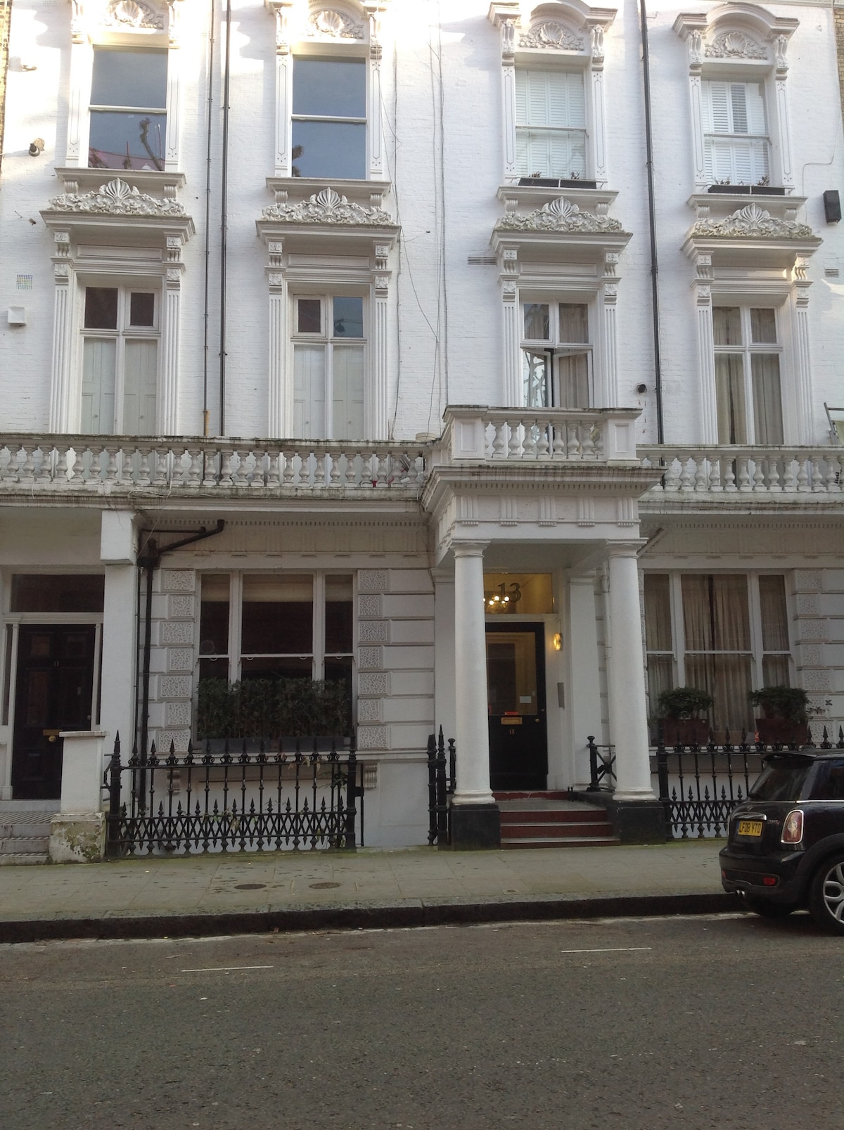 A Victorian-style building is presented, featuring ornate architectural details and a decorative facade. Tall windows are visible, framed by elegant moldings. A small staircase leads to the entrance, flanked by planters at either side. A wrought-iron railing borders the front area.