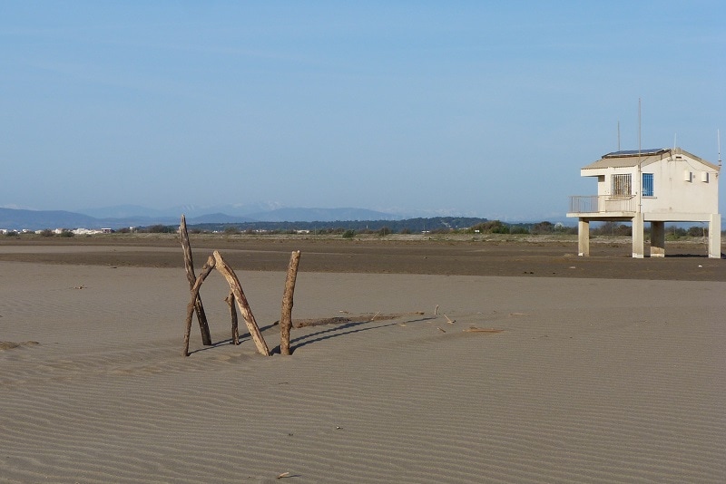 A sandy landscape features wooden structures shaped by weathering, set against the backdrop of a distant building perched on stilts. Soft waves of sand create a textured surface under a clear blue sky, contributing to a serene beach atmosphere.
