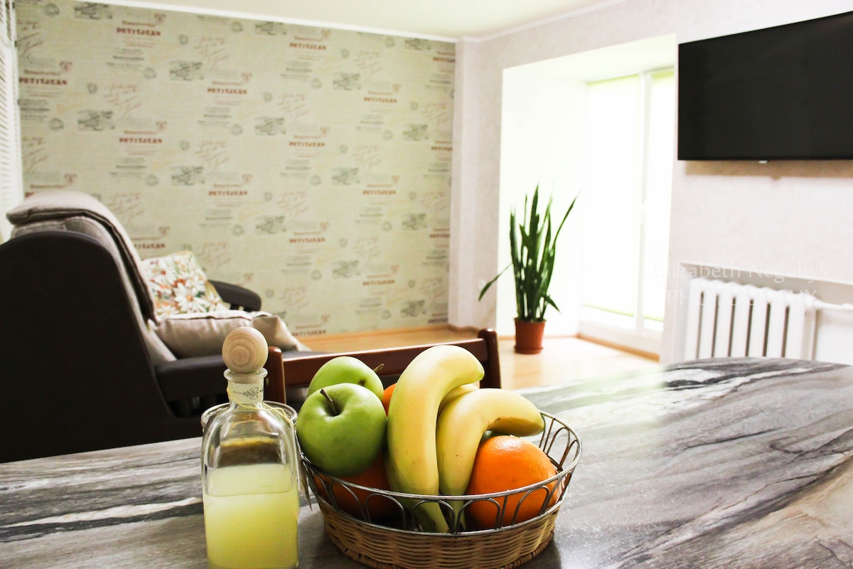 A bowl filled with fresh fruit, including green apples, bananas, and an orange, is placed on a marble countertop. The living area features a comfortable sofa and a wall-mounted television, with a large window allowing natural light to fill the space.