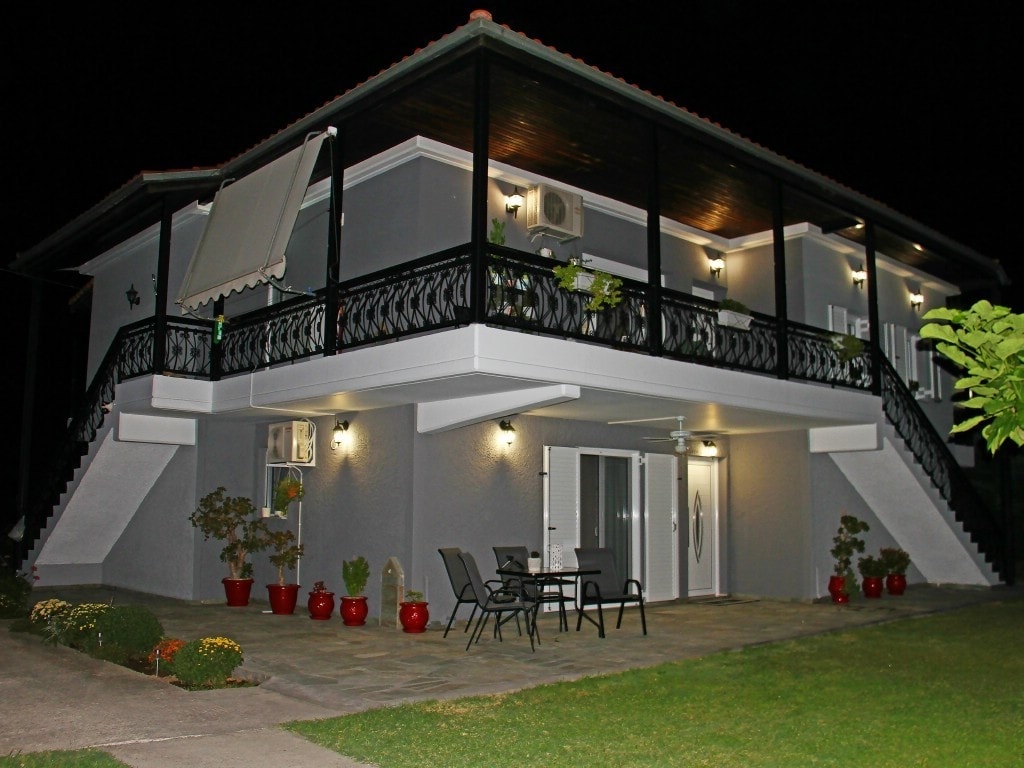 A modern two-story building is illuminated at night, featuring a spacious balcony with decorative railings. Outdoor seating is visible on the patio, surrounded by a well-maintained garden with potted plants. Soft lighting accentuates the structure's elegant design against a dark sky.