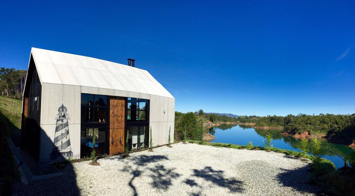 The modern chalet features a unique design with a striking combination of concrete and wooden elements. Large windows reflect the natural surroundings, and a gravel area provides access to picturesque lake views. Lush greenery borders the property, enhancing the serene atmosphere.