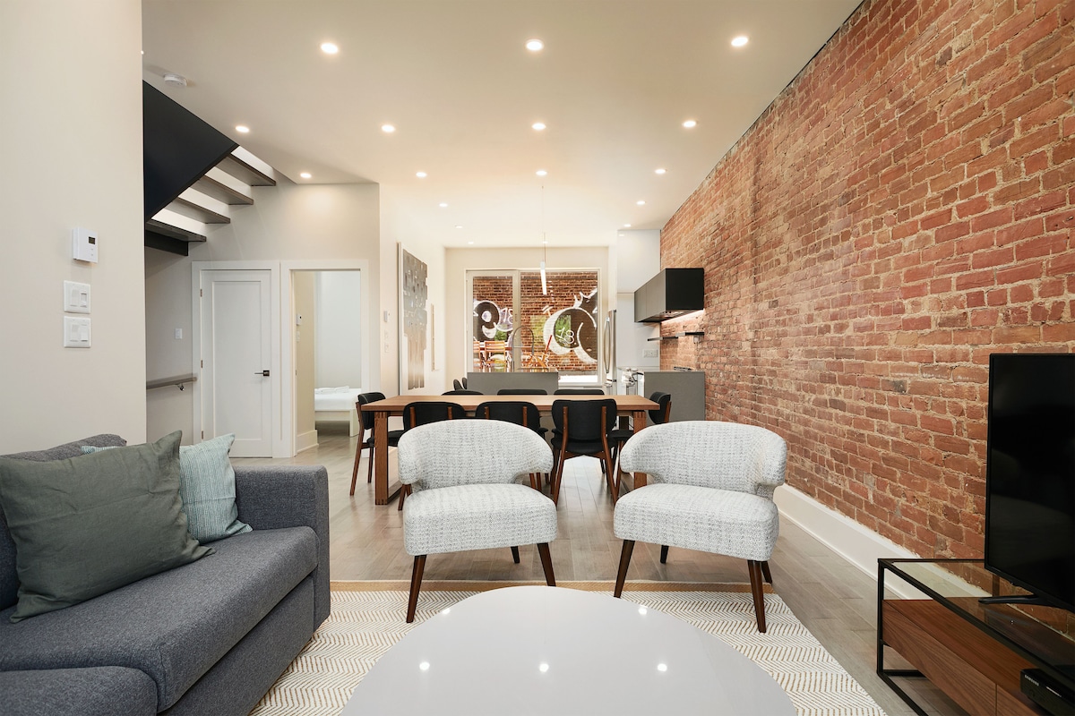 The common area features exposed brick walls and modern furnishings. Two light grey armchairs are set opposite a low coffee table, with a grey sofa nearby. A dining area is visible in the background, accompanied by sleek cabinetry and a round light fixture above the table.