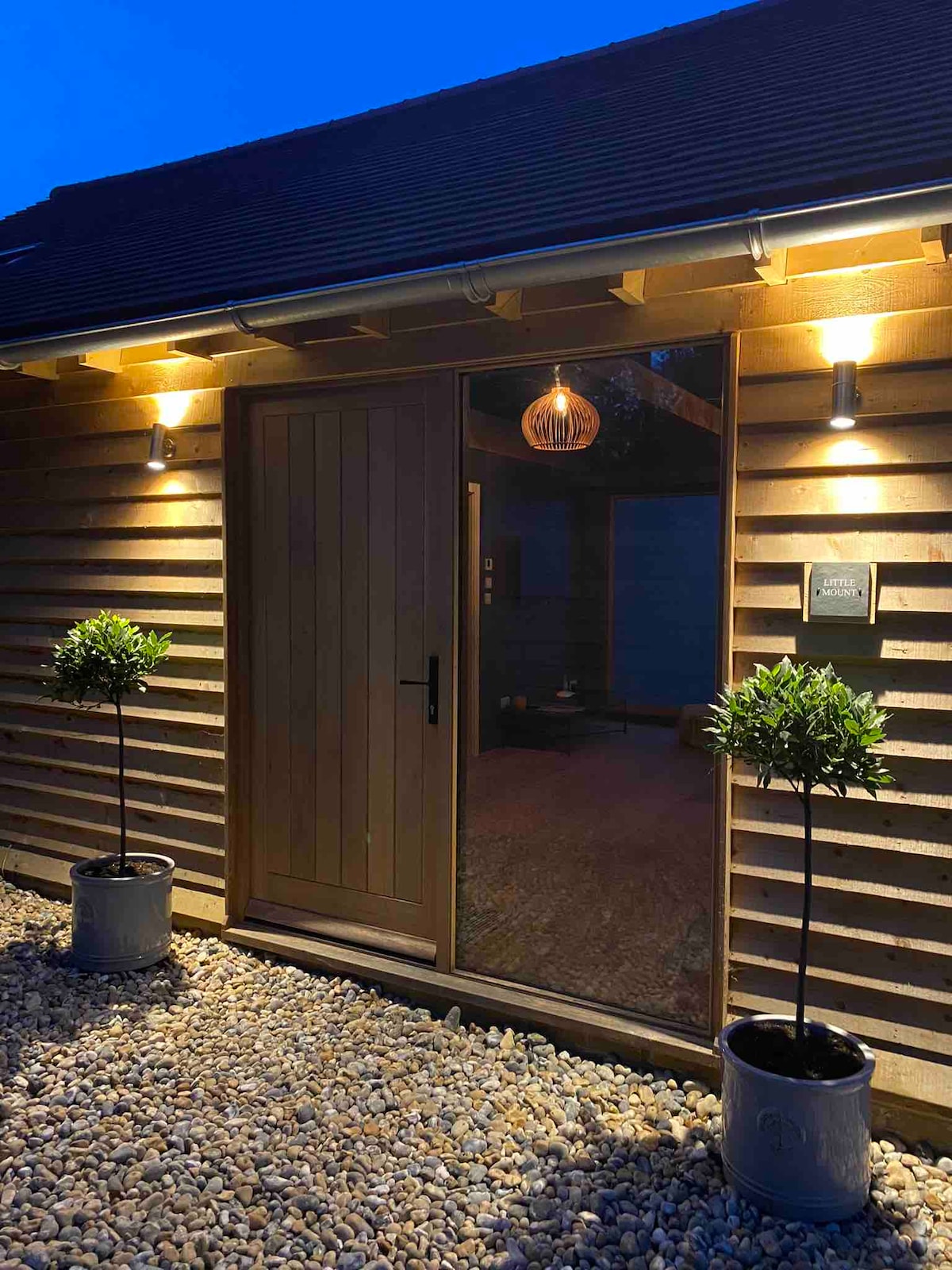 The entrance of a cottage is framed by two potted topiary plants on either side. Soft lighting illuminates the wooden door and the textured siding of the building, creating a welcoming atmosphere. A decorative lantern hangs above the door, complementing the serene evening setting.