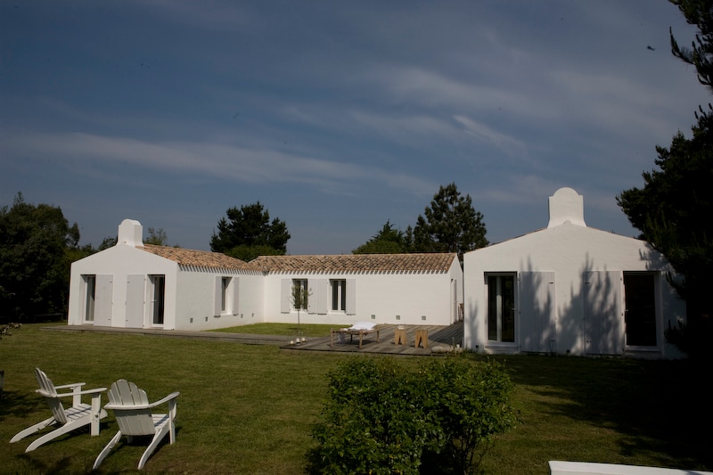 A spacious white house with a modern design is situated in a grassy area. The building features multiple sections with a tiled roof, large windows, and outdoor seating. Surrounding greenery enhances the tranquil setting under a clear blue sky.