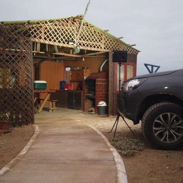 A pathway leads to an open structure featuring wooden lattices. Inside, cabinets and counters are visible, suggesting a functional space. A vehicle is parked nearby, providing easy access. Natural surroundings contribute to a serene atmosphere.
