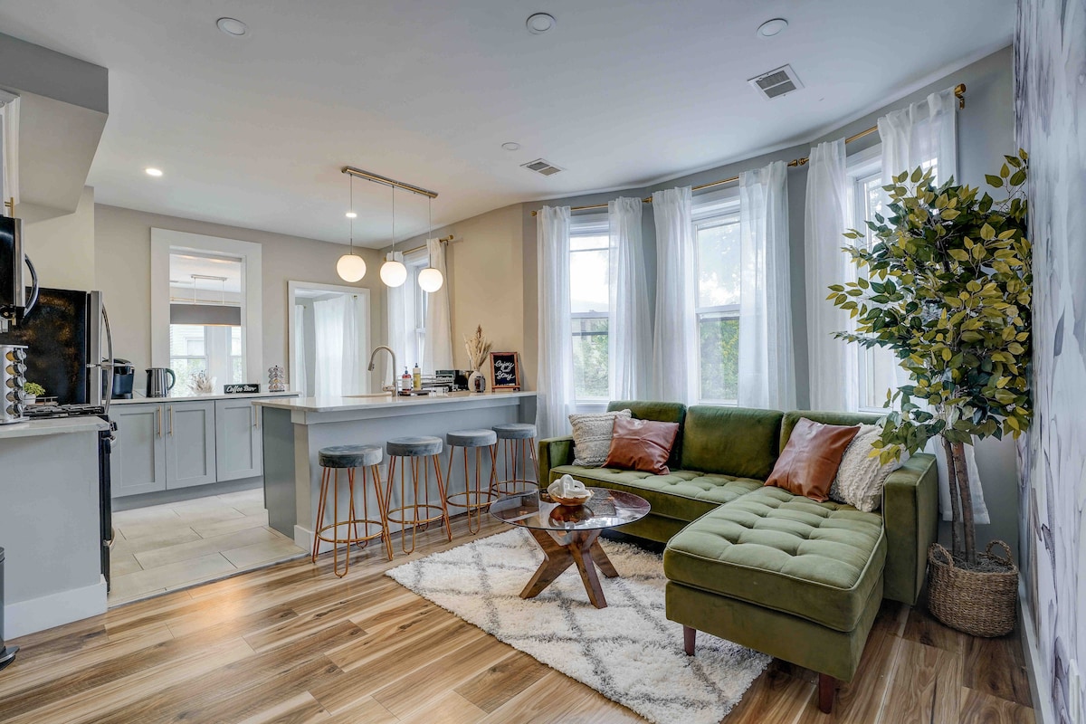 A modern living space features a plush green sectional sofa adorned with decorative cushions. A wooden coffee table sits atop a soft area rug, while stylish bar stools are positioned at a sleek kitchen island. Abundant natural light filters through large windows, enhancing the inviting atmosphere.
