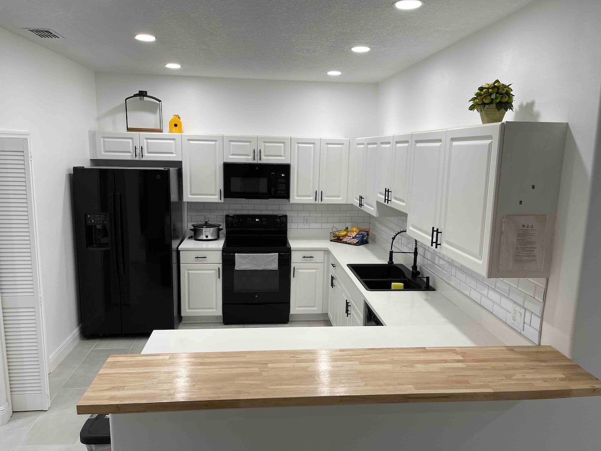 A modern kitchen features white cabinetry contrasted with a black refrigerator and stove. A spacious countertop provides room for meal preparation. A clean backsplash with square tiles complements the sleek design, while soft overhead lighting enhances the bright atmosphere. A small plant decorates the countertop.