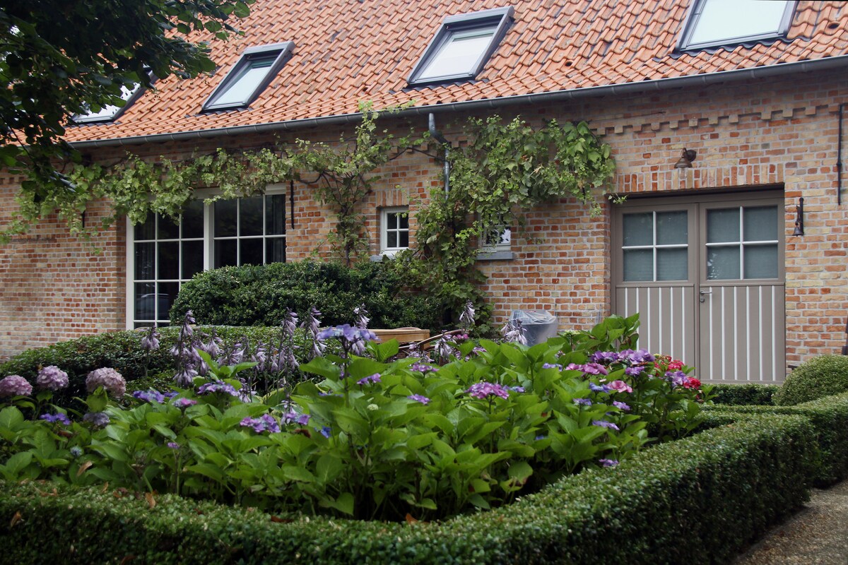 A charming exterior view of the vacation home is featured, showcasing a well-maintained garden with lush green hedges and vibrant flowering plants. The brick facade is complemented by a traditional roof with skylights, while a wooden door adds to the welcoming entryway.