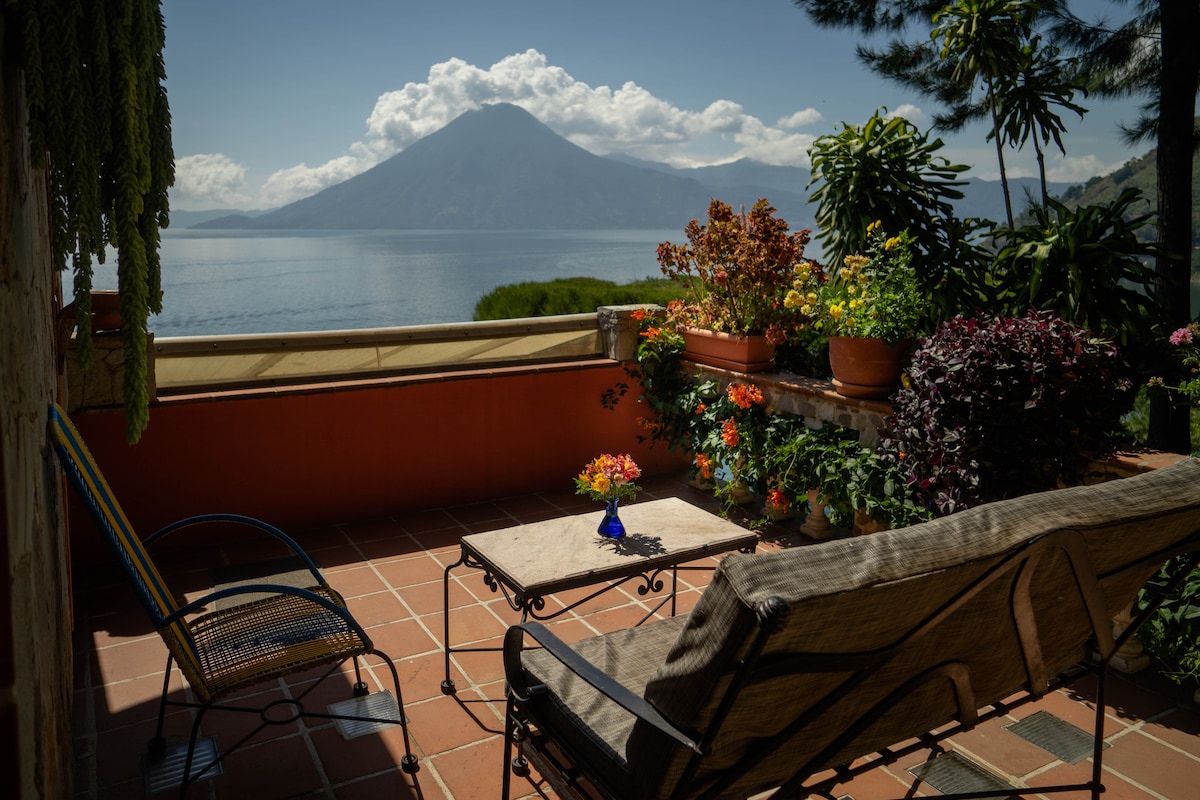 A serene private balcony is adorned with potted plants and a small vase of flowers on the table. Comfortable seating overlooks a stunning view of Lake Atitlan and the surrounding mountains under a clear sky.