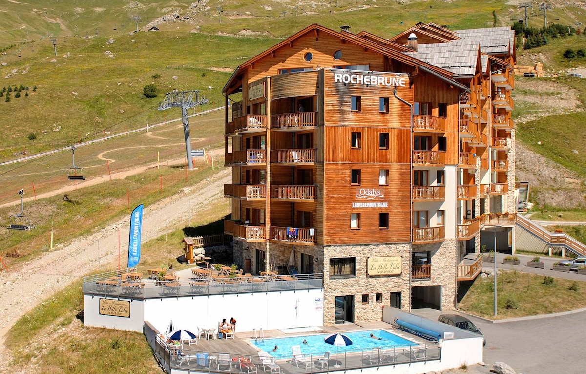 A multi-story building is situated at the foot of ski slopes, featuring wooden accents and balconies. The outdoor heated swimming pool is visible at the base, accompanied by umbrellas and lounge chairs. Surrounding mountains provide a scenic backdrop, completing the alpine atmosphere.