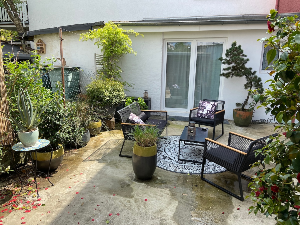 An inviting outdoor space features comfortable black wicker chairs arranged around a small table. Lush greenery and decorative plants are present in large pots, with a patterned rug placed on the ground. Natural light illuminates the area, enhancing the tranquil atmosphere.