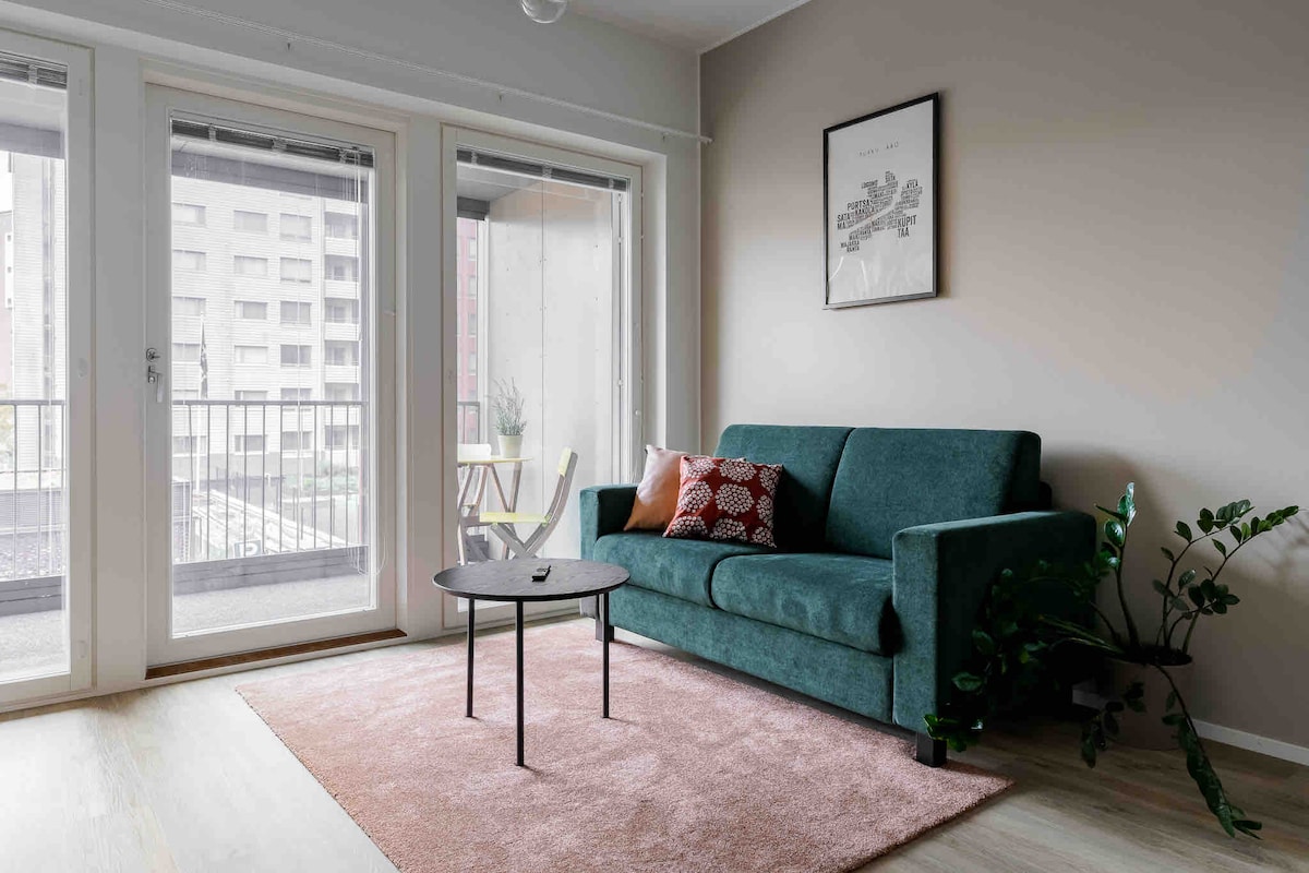 A cozy living area features a green sofa adorned with decorative cushions, positioned adjacent to large glass doors that lead to a balcony. A circular black coffee table rests on a soft area rug, with natural light filtering in from outside.