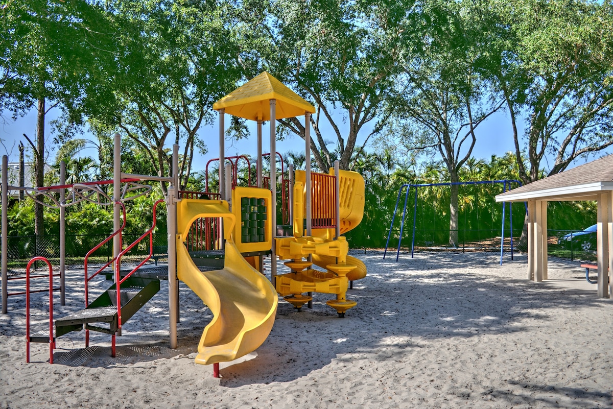 A vibrant playground area features a yellow play structure with a slide and climbing equipment, surrounded by swings. Sandy ground is evident, providing a safe play surface. Lush green trees frame the area, offering shade and a natural setting for family fun.