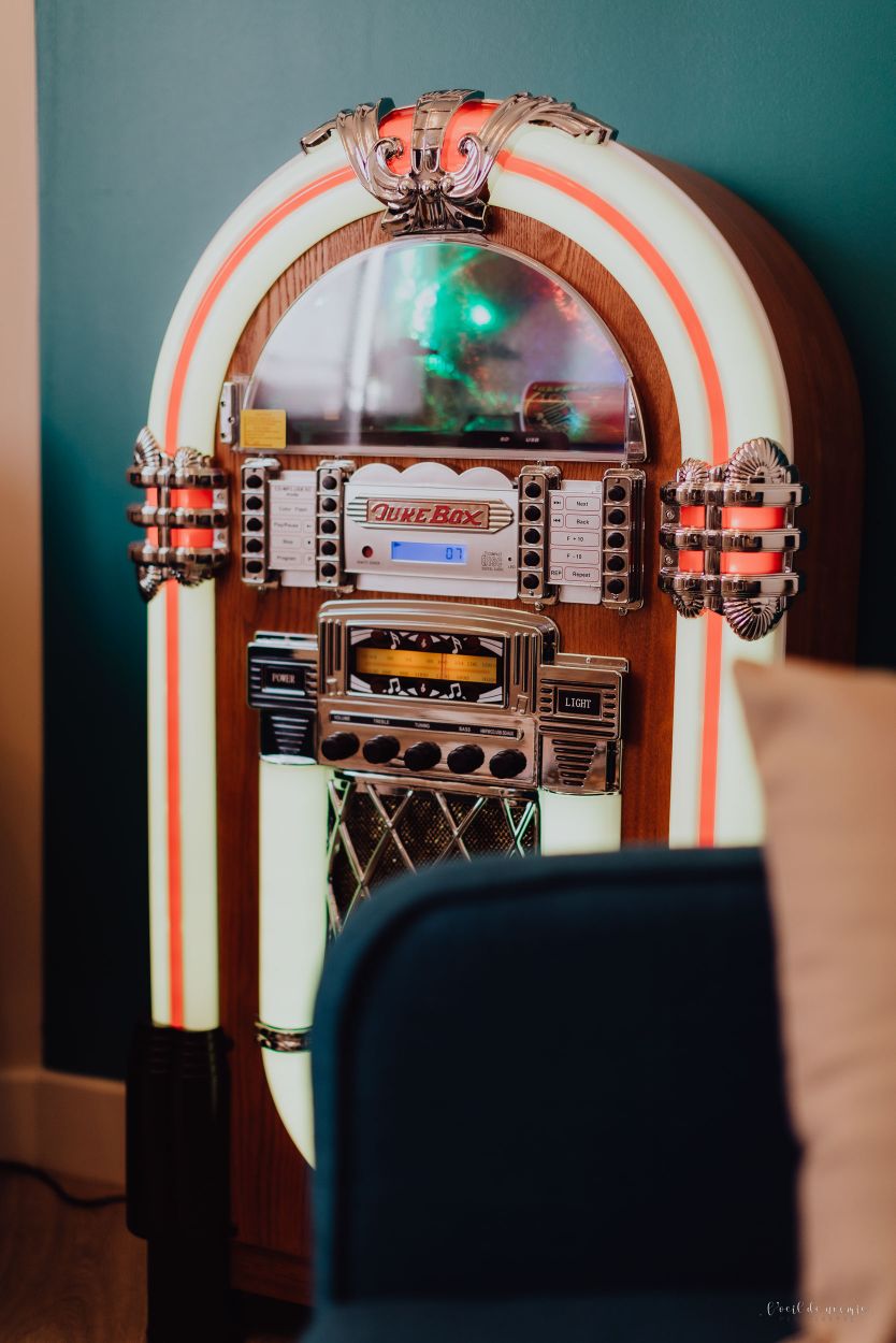 A vintage jukebox features a polished wooden frame with chrome detailing and illuminated accents in soft colors. The glossy front showcases various buttons and a display, evoking a nostalgic ambiance within the living space.