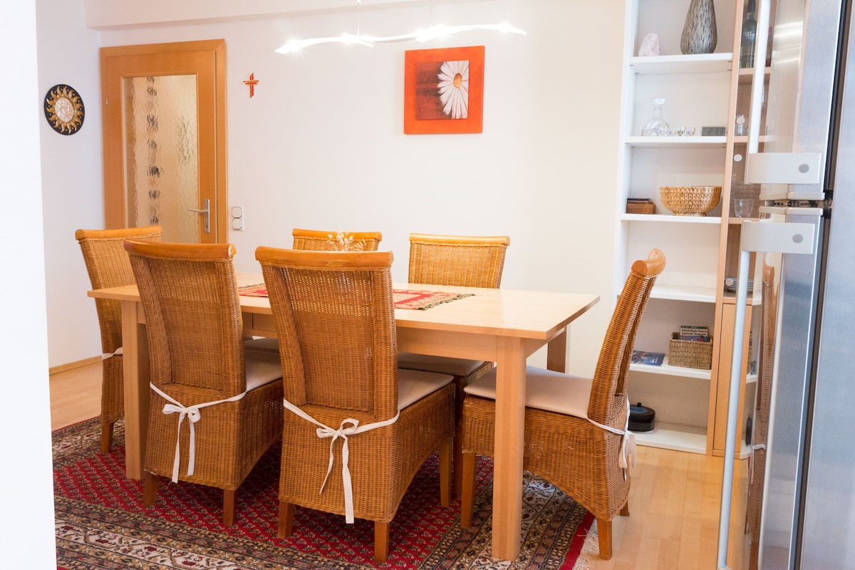A dining area is shown with a wooden table surrounded by six woven chairs. A decorative table setting is present, and a stylish light fixture hangs above. A patterned rug covers the floor, and shelves with various decorative items are visible in the background.