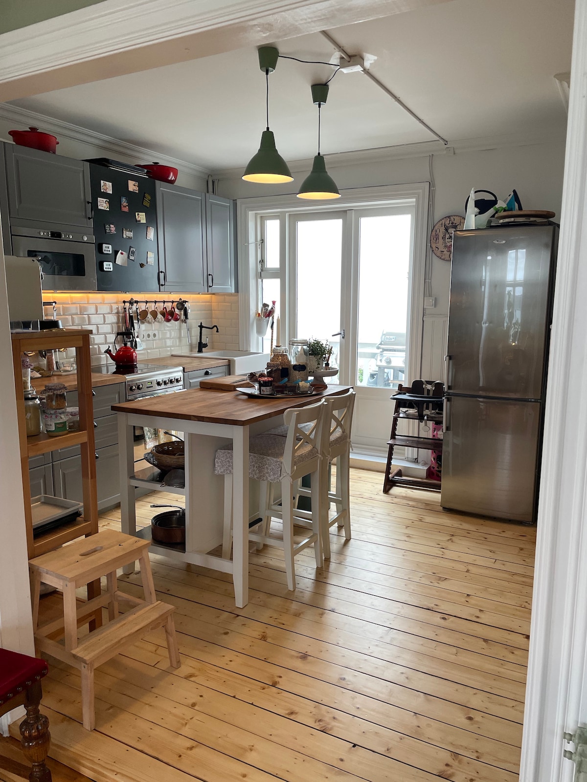 The modern kitchen features gray cabinetry and wooden floors, illuminated by green pendant lights. A small dining area is created with a breakfast bar and stools. The kitchen is equipped with stainless steel appliances and has large windows allowing natural light to fill the space.