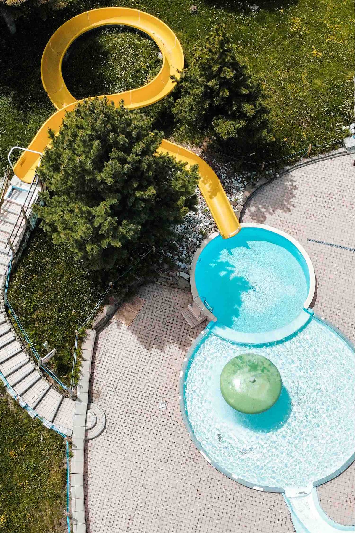 An aerial view showcases a vibrant yellow waterslide leading into a circular swimming pool. The pool is surrounded by green grass and trees, offering a refreshing atmosphere for relaxation and play. The bright colors contrast with the surrounding natural landscape.